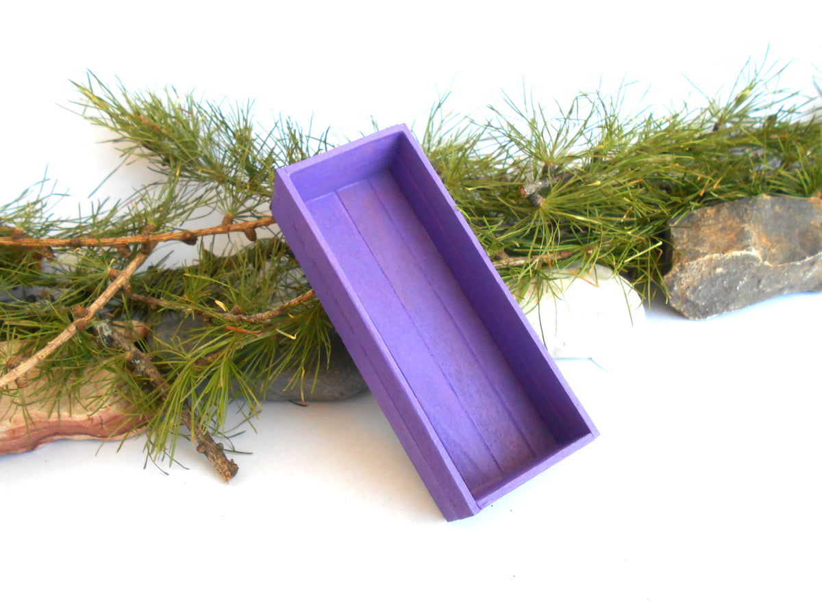 A purple rectangular wooden box with an open lid, styled on a white background with green pine branches and small rocks.