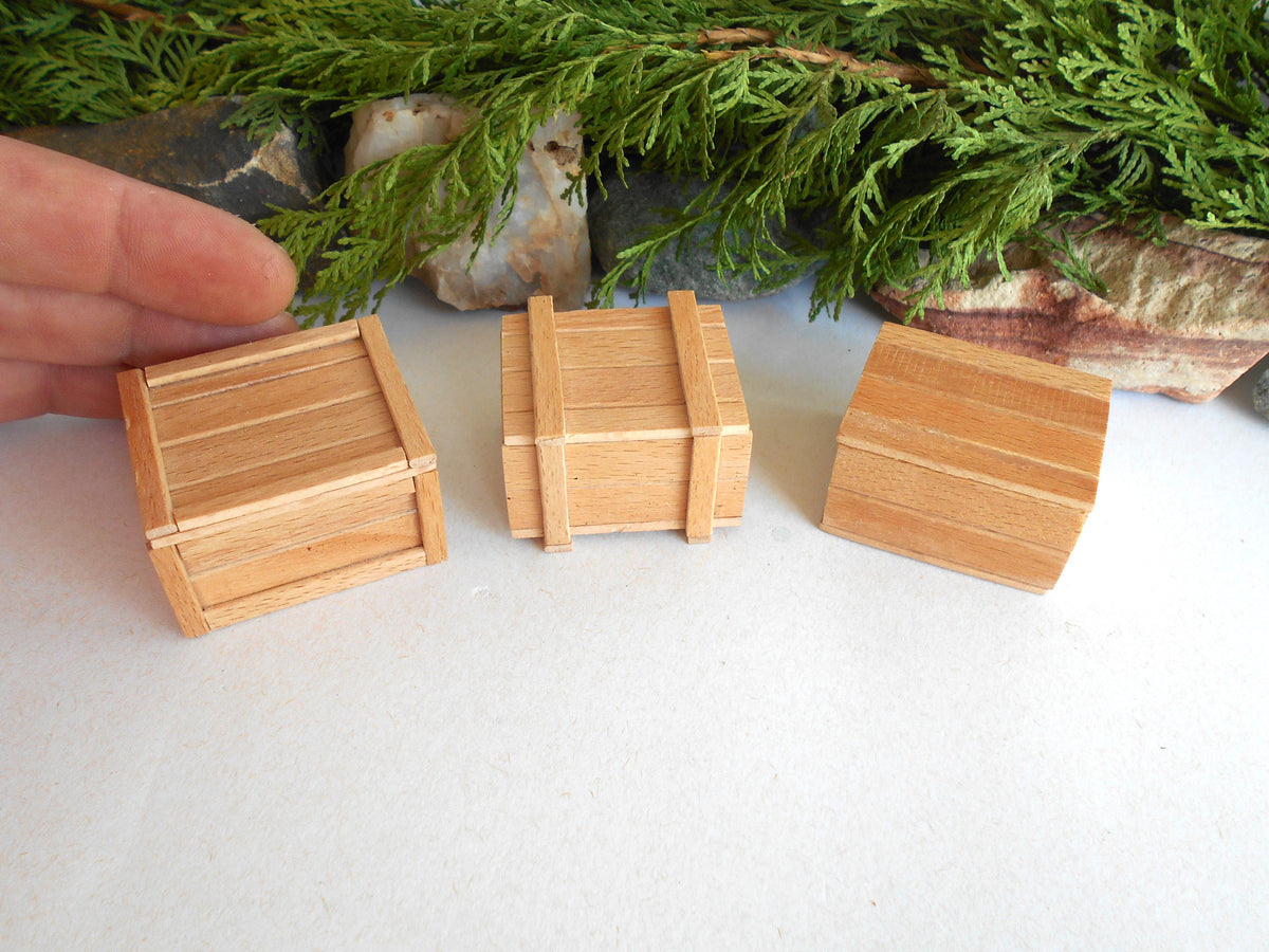 Three miniature light wood boxes of different shapes and sizes displayed on a white surface, with a hand holding one box on the left and green foliage in the background.