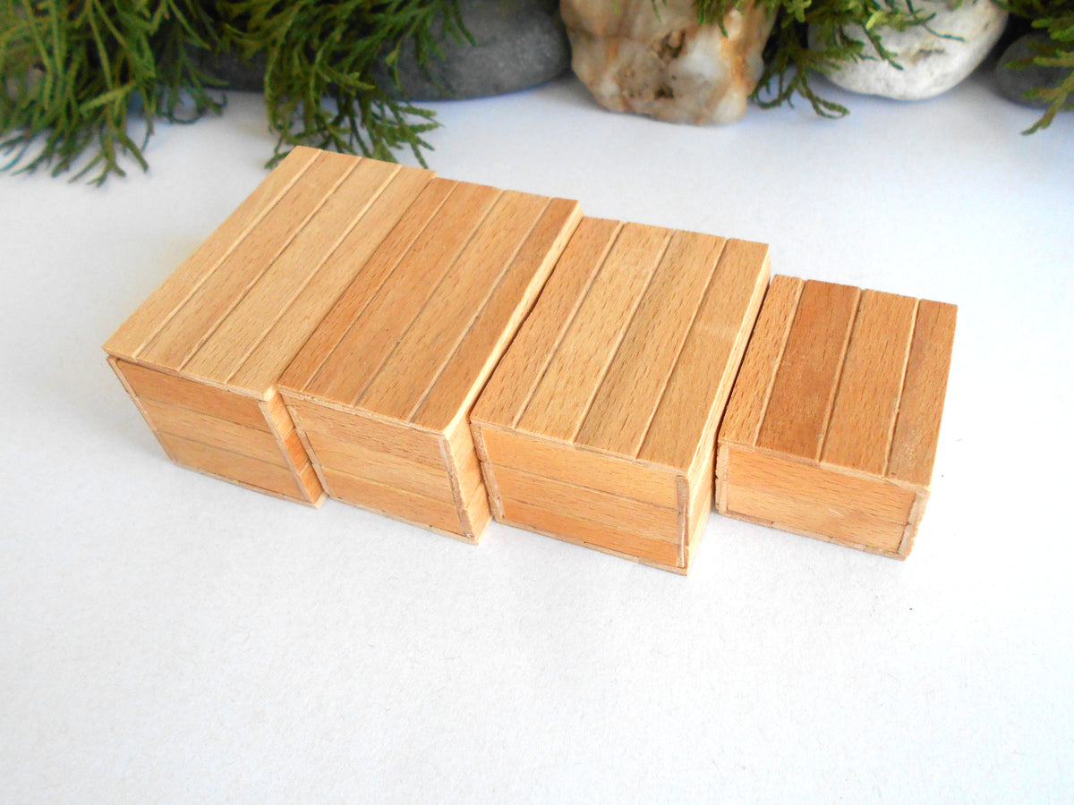 Four small rectangular wooden boxes arranged in a staggered row on a white surface, with decorative greenery and stones blurred in the background.