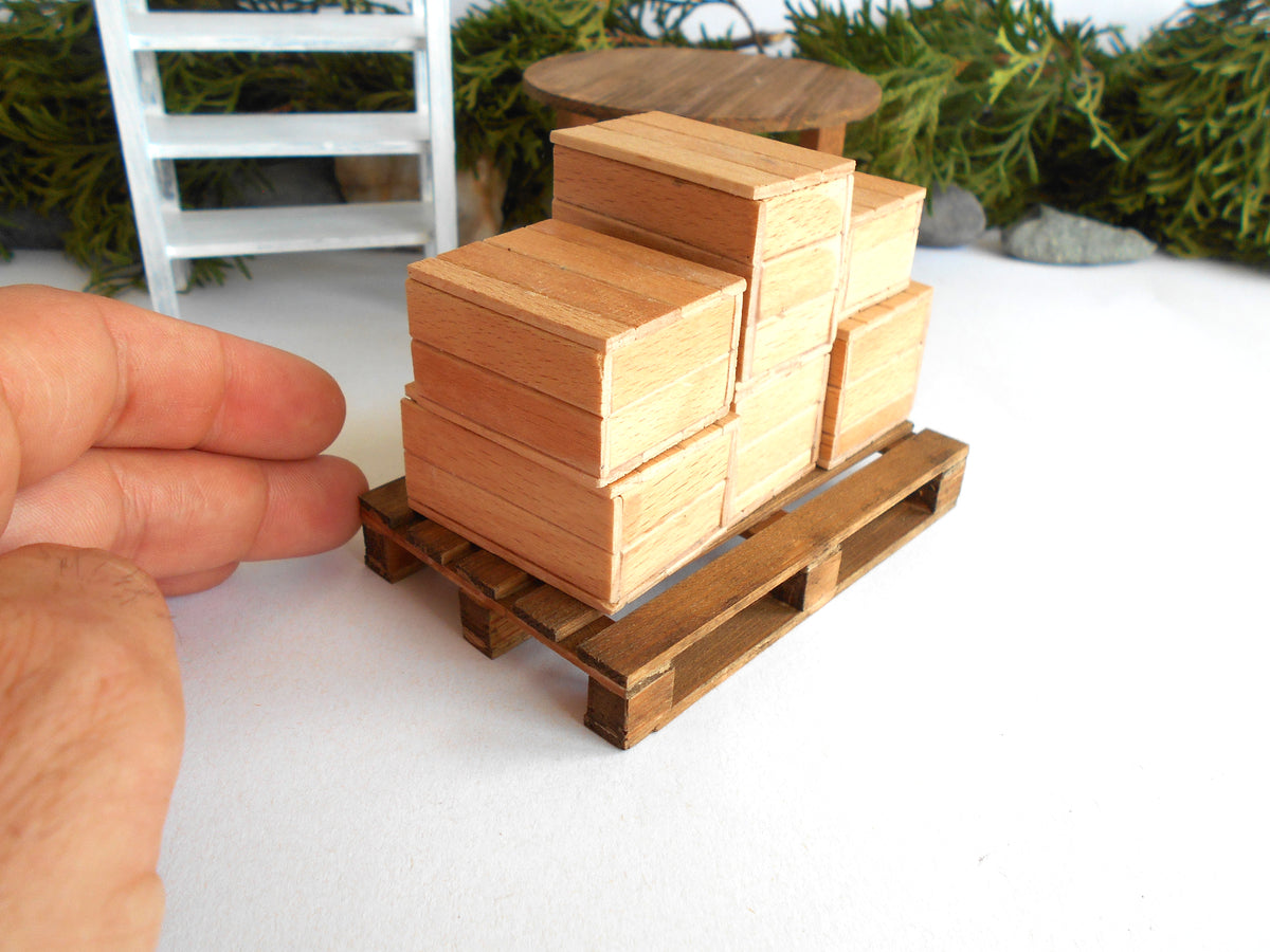 A miniature wooden pallet toy with stacked wooden box blocks, shown next to a human hand for scale on a white surface.