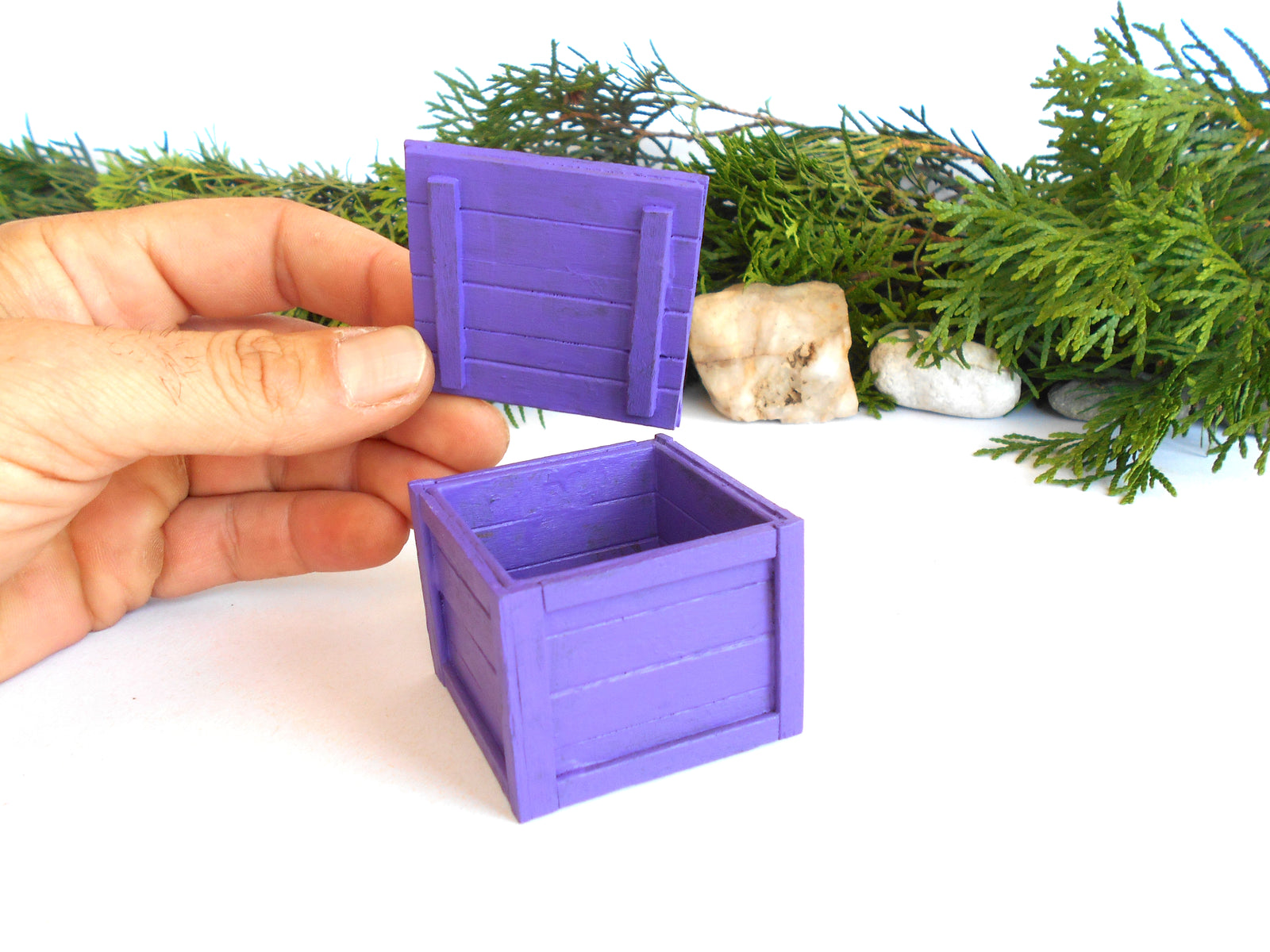 A hand holding a small purple wooden crate lid next to an open matching purple crate, with green foliage and small stones in the background on a white surface.