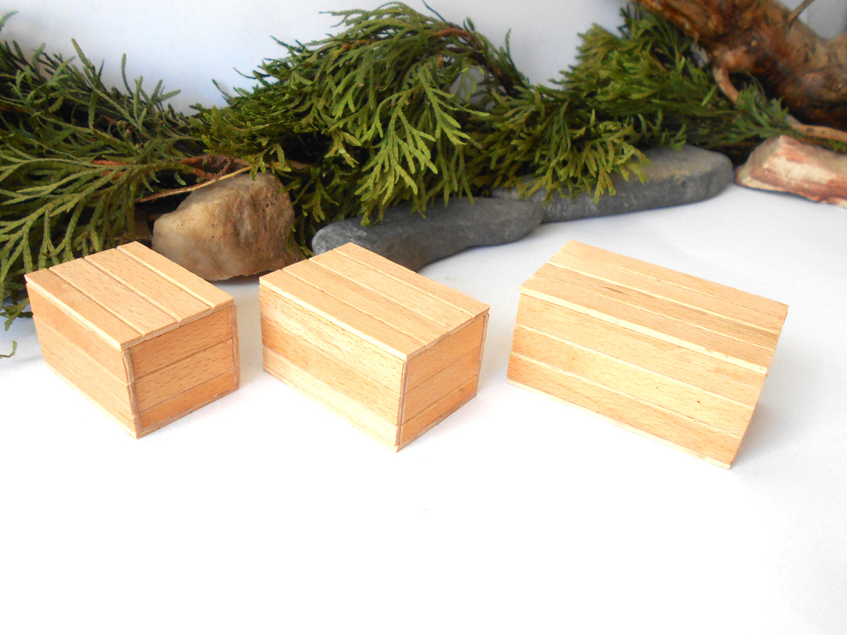 Three small rectangular wooden boxes with a natural light wood finish, arranged on a white surface with green pine branches and stones in the background.