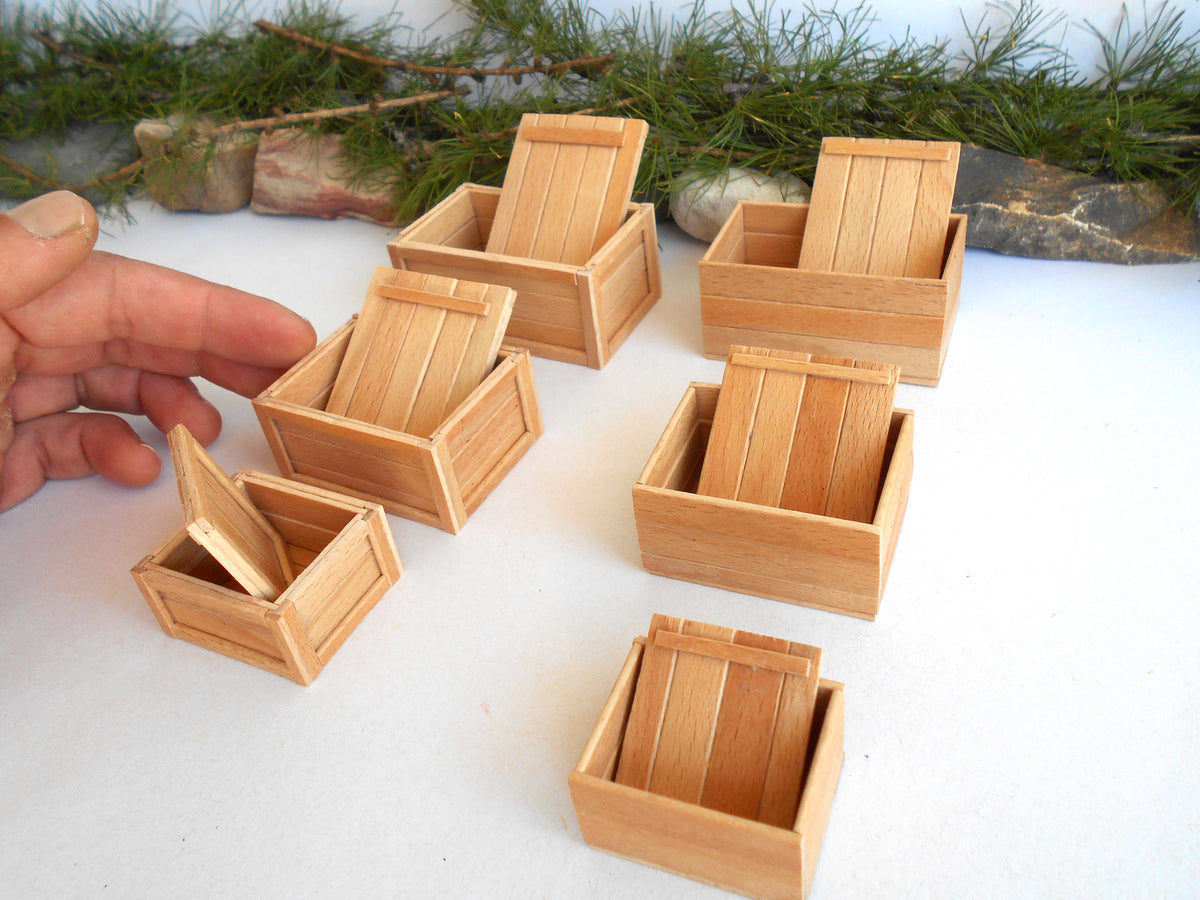 Six miniature light wood boxes with hinged lids are arranged on a white surface, with a hand pointing to one of the boxes on the left. Pine branches and small stones are visible in the background.