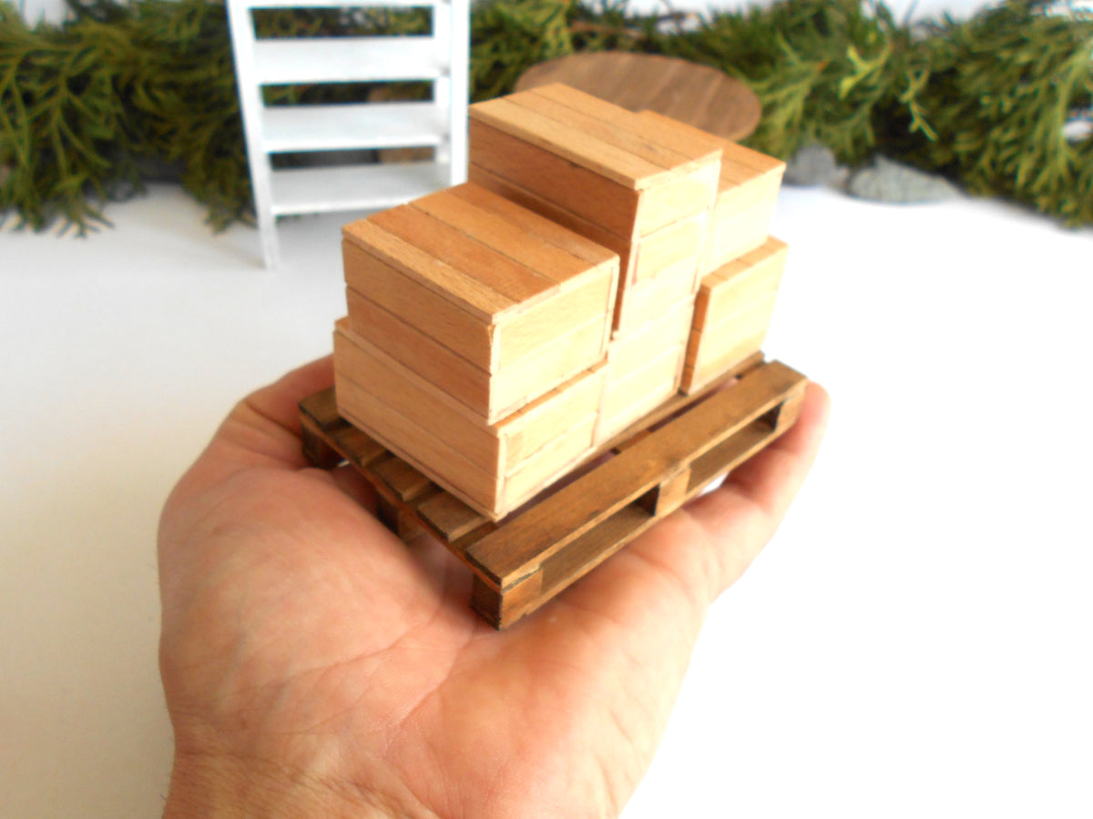 A hand holding a miniature wooden pallet with stacked wooden boxes, shown in a close-up shot with a white surface and blurred greenery in the background.