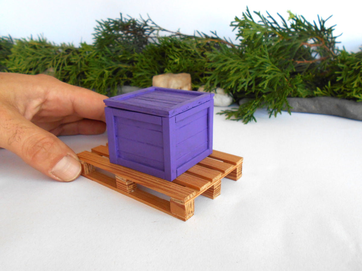 A miniature purple wooden crate box resting on a small wooden pallet, with a human hand holding it for scale against a white surface with green foliage in the background.