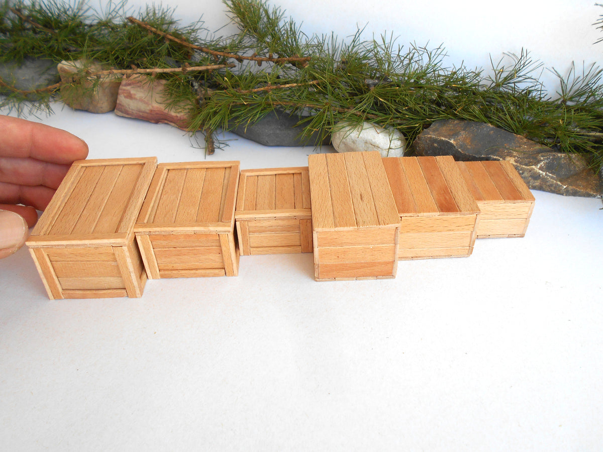 A set of miniature light wood crates arranged in a row on a white surface, with a hand holding one crate on the left and pine branches and stones in the background.