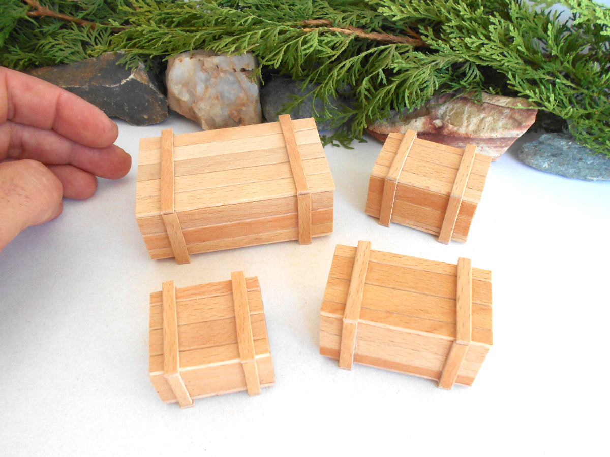 Four miniature wooden crates with light-colored wood grain and darker brown bands, displayed on a white surface with a hand on the left for scale and green foliage in the background.