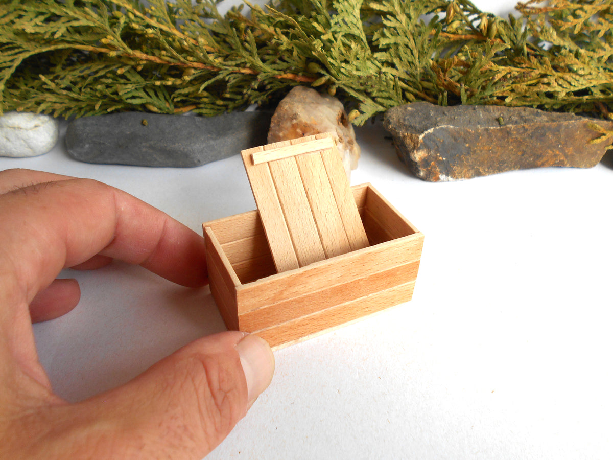 A miniature wooden box with a hinged lid, held by a hand to show its small scale, set against a white background with decorative greenery and rocks.