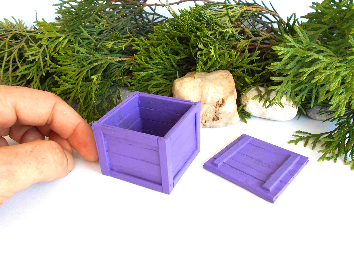 A miniature purple wooden crate box with its lid removed, held by a hand on a white surface with green foliage and small stones in the background.