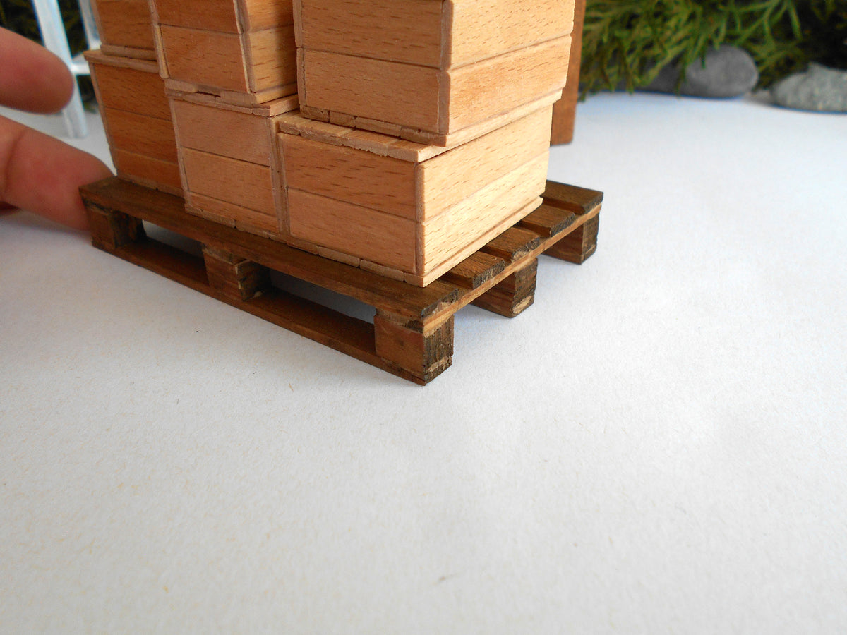 A miniature wooden pallet model with stacked wooden block cargo on top, held by a person's finger on the left, set against a white surface with a blurred background.