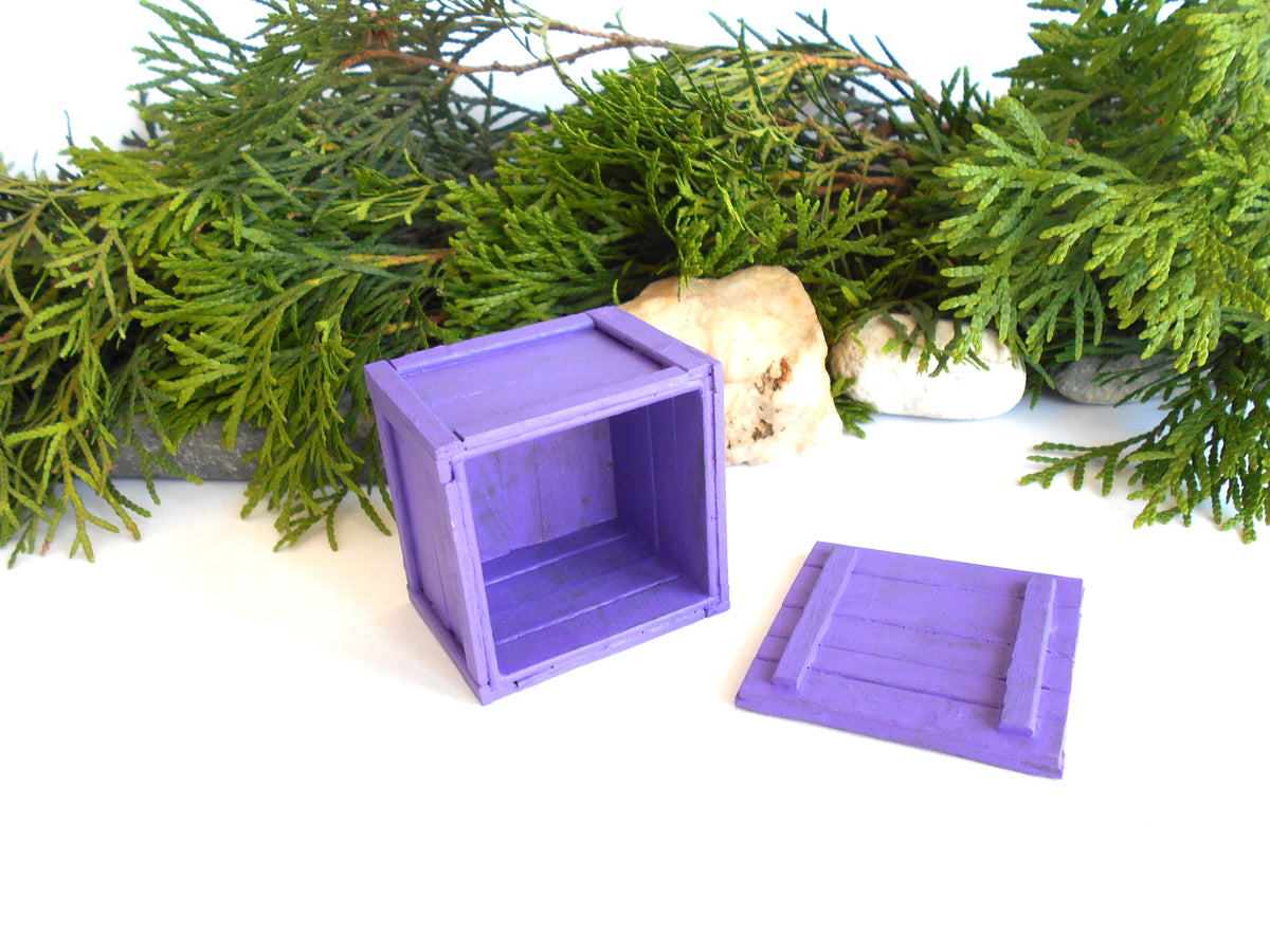 A small purple wooden crate with its lid placed beside it on a white surface, styled with green evergreen branches and small stones in the background.