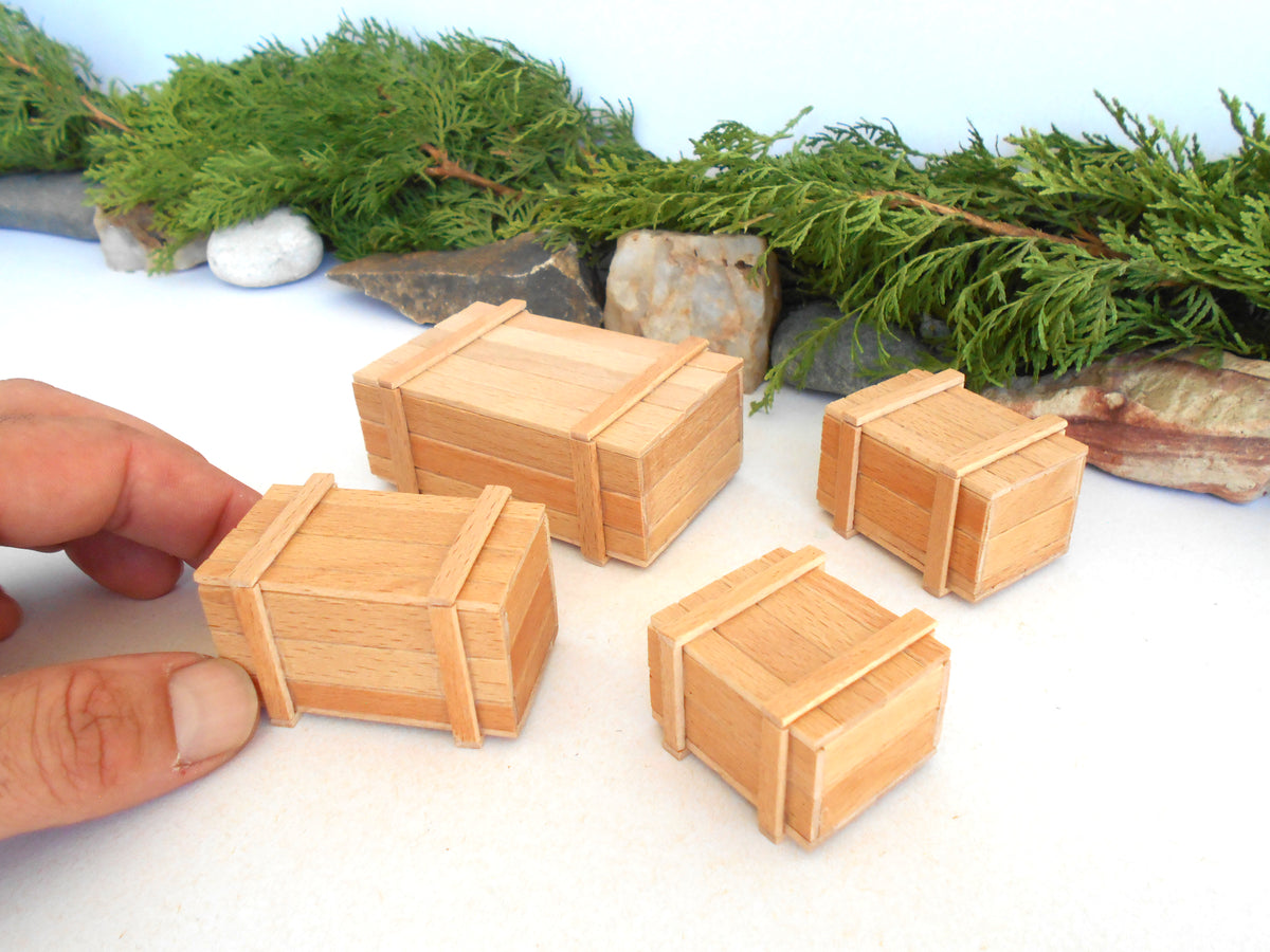 Four miniature wooden crates with light-colored wood grain and darker brown bands, displayed on a white surface with a hand on the left for scale and green foliage in the background.