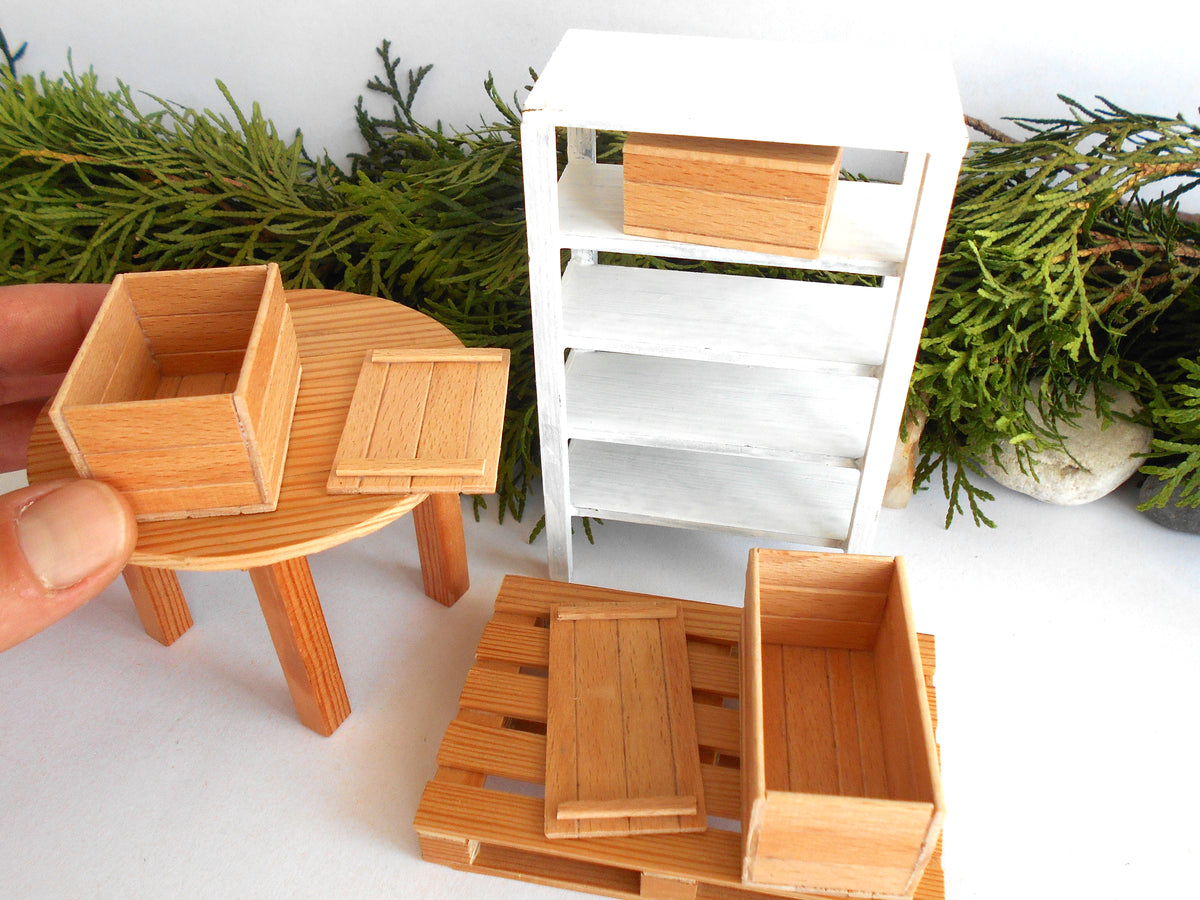 A miniature dollhouse furniture set featuring a round table, shelving unit, and wooden crates, with a hand holding one of the crates on the left.