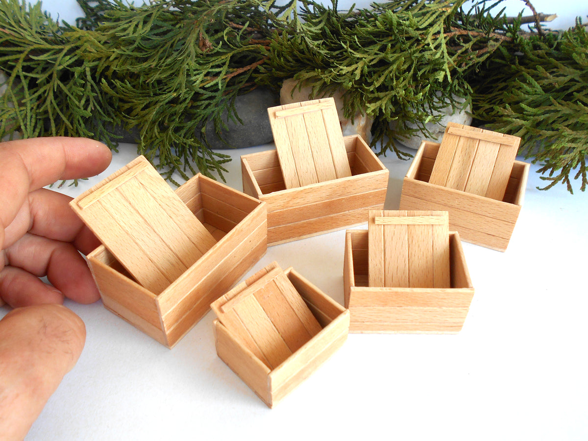 A set of miniature light wood boxes with hinged lids, shown with a hand holding one box to demonstrate scale, against a white background with green foliage at the top.