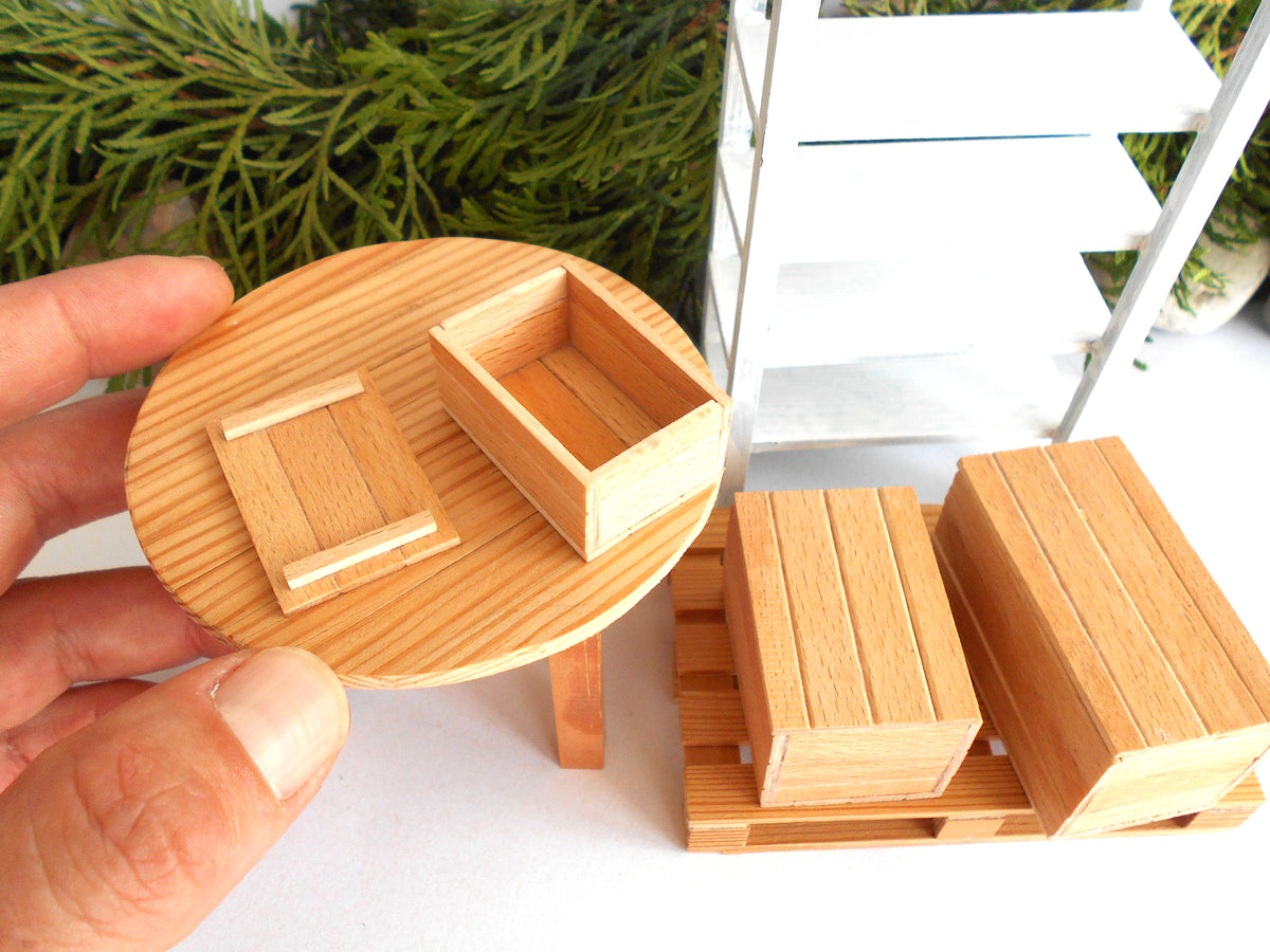 A miniature wooden dollhouse furniture set including a round table with a crate, a ladder, and storage boxes on pallets, shown with a hand holding a crate on the table.