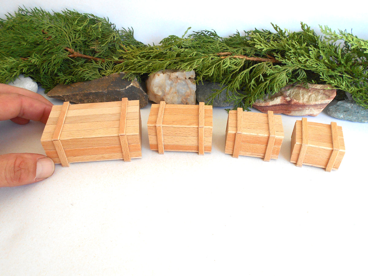 Four miniature light wood toy chests of varying sizes arranged in a row on a white surface, with a hand holding the leftmost chest and green foliage and stones in the background.