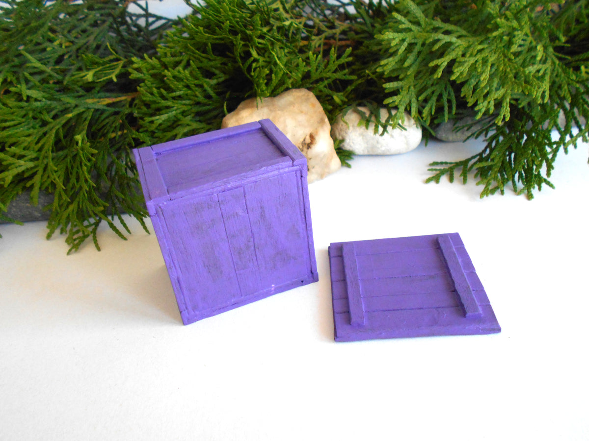 A small purple wooden crate with its lid placed beside it on a white surface, styled with green foliage and small stones in the background.