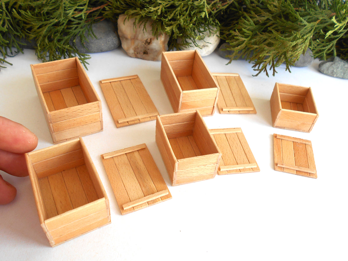 A set of miniature light wood crates and boxes, including some open storage bins and flat slatted lids, arranged on a white surface with green foliage and small stones in the background. A person's fingers are visible on the left holding one of the crates to show scale.
