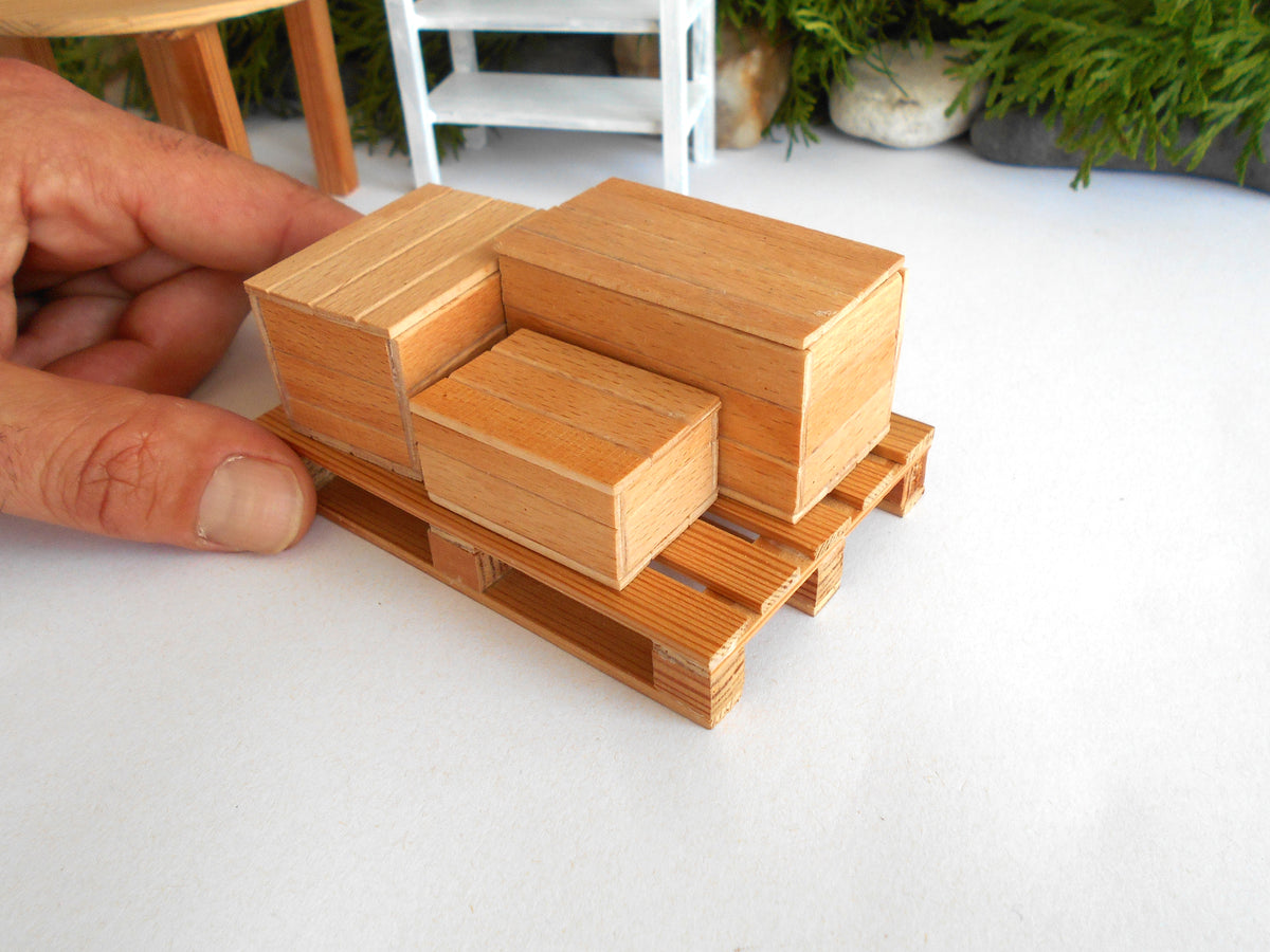 A close-up of a miniature wooden pallet and stacked storage boxes, with a hand holding the side of the pallet to show scale, on a white surface.