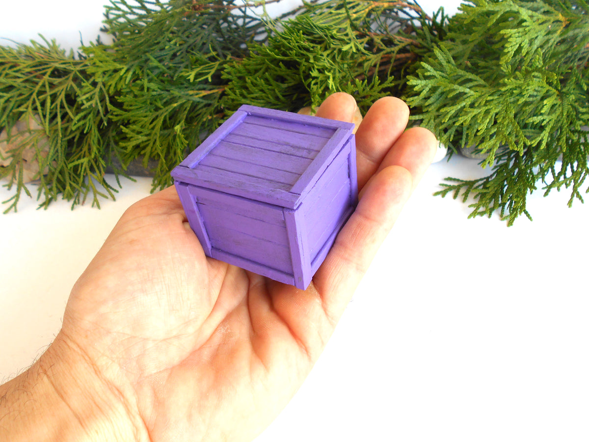 A miniature purple wooden crate model held in a person's hand, with green pine branches in the background.