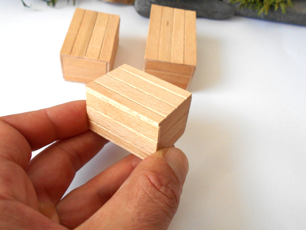 A hand holding a small rectangular wooden block with visible wood grain, with two similar blocks in the background on a white surface.