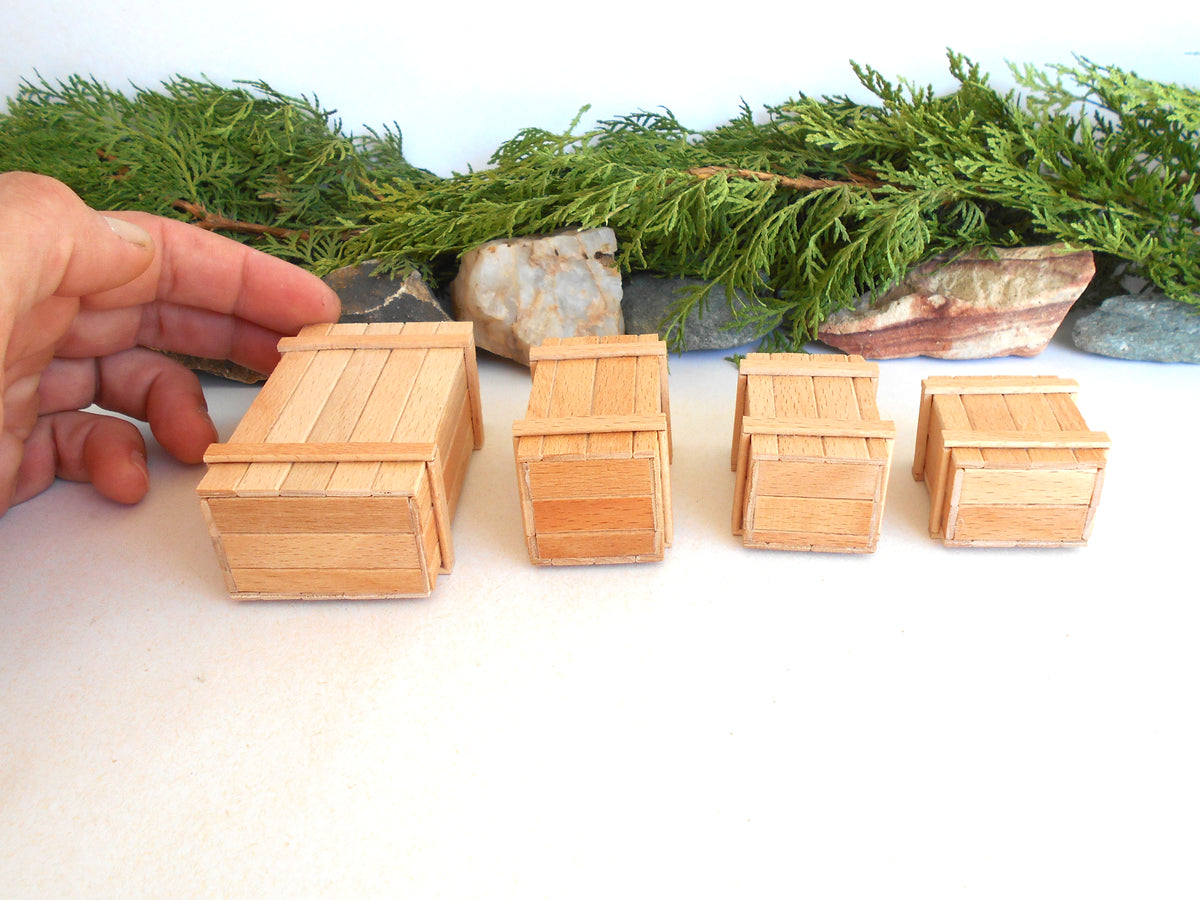 Four miniature light wood crates of varying sizes arranged on a white surface, with a hand pointing to the leftmost crate and green foliage in the background.