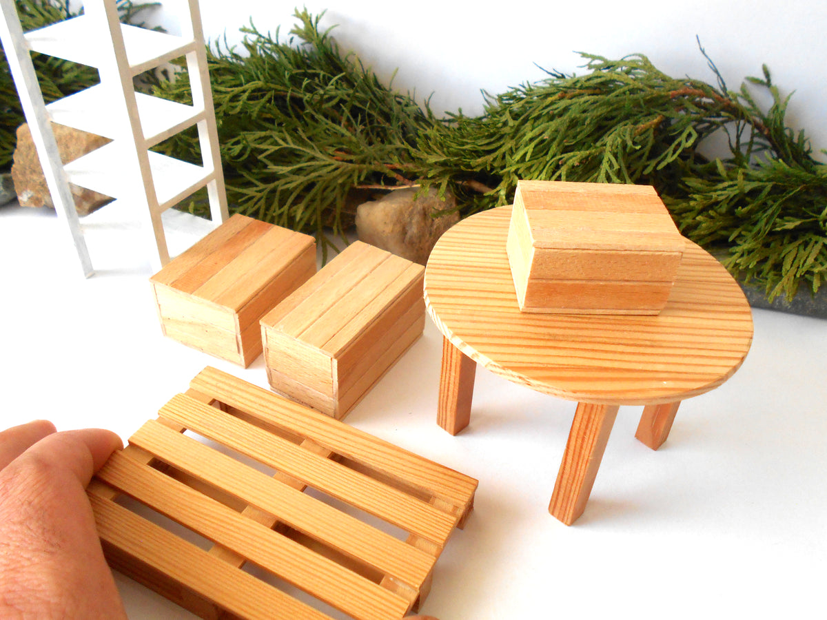 A miniature wooden dollhouse furniture set including a round table with legs, two rectangular blocks, and a slatted base, shown with a hand holding one piece and green foliage in the background.