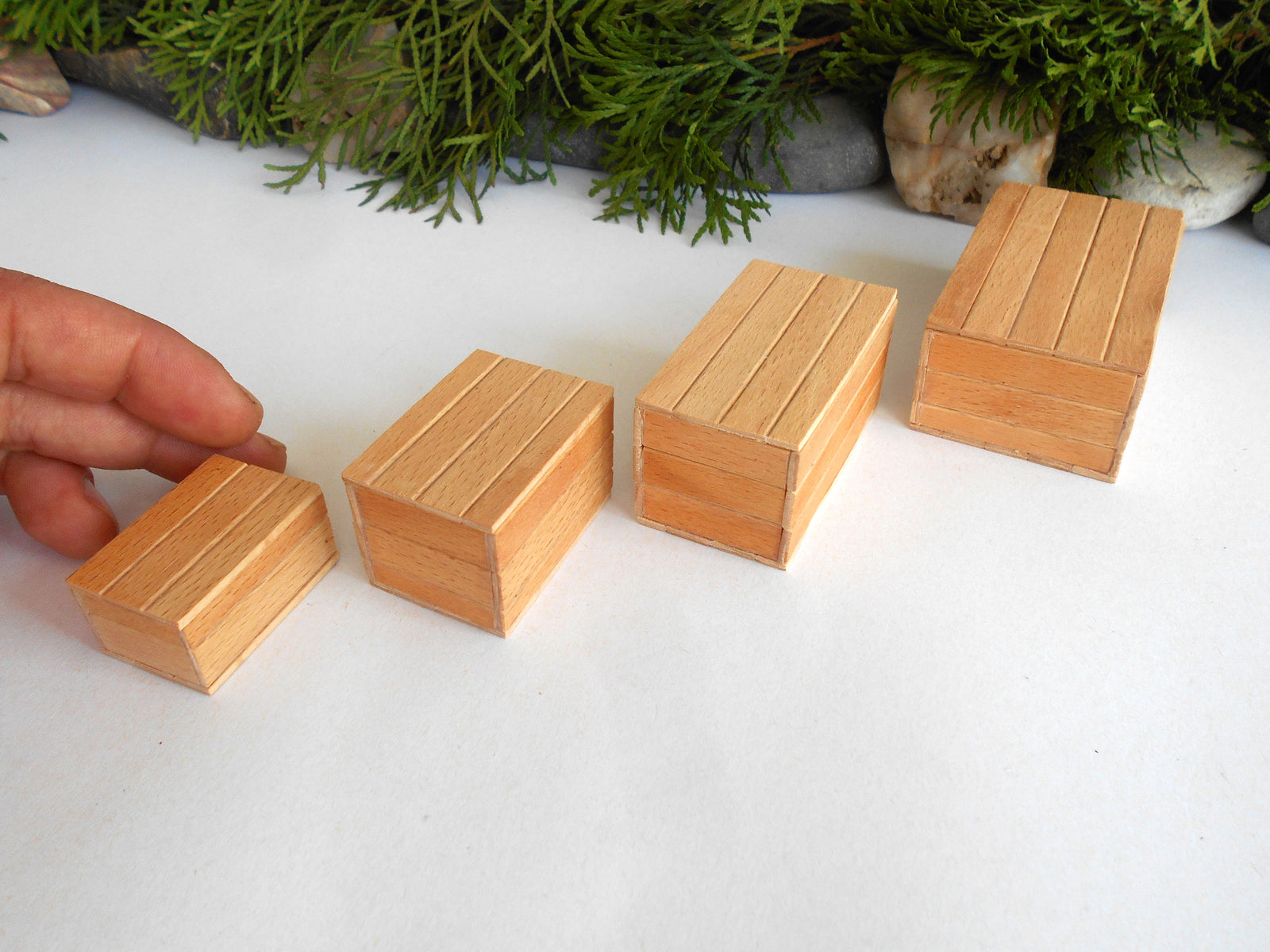 Four small rectangular wooden boxes arranged in ascending size order on a white surface, with a hand touching the smallest box. Green foliage decor is visible in the background.