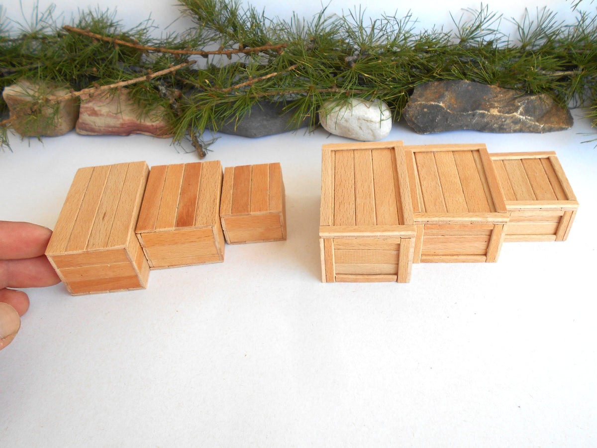 A set of miniature light wood crates in different sizes, with a hand holding one of the smaller crates on the left, set against a white background with decorative pine branches and stones at the top.