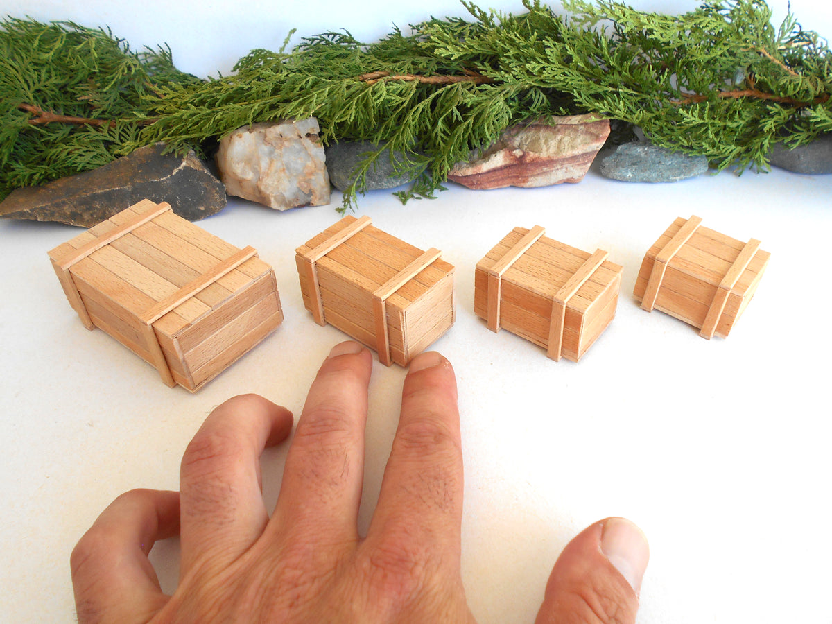 Four miniature light wood boxes of varying sizes arranged on a white surface, with a person's hand visible for scale and greenery and rocks along the top edge.