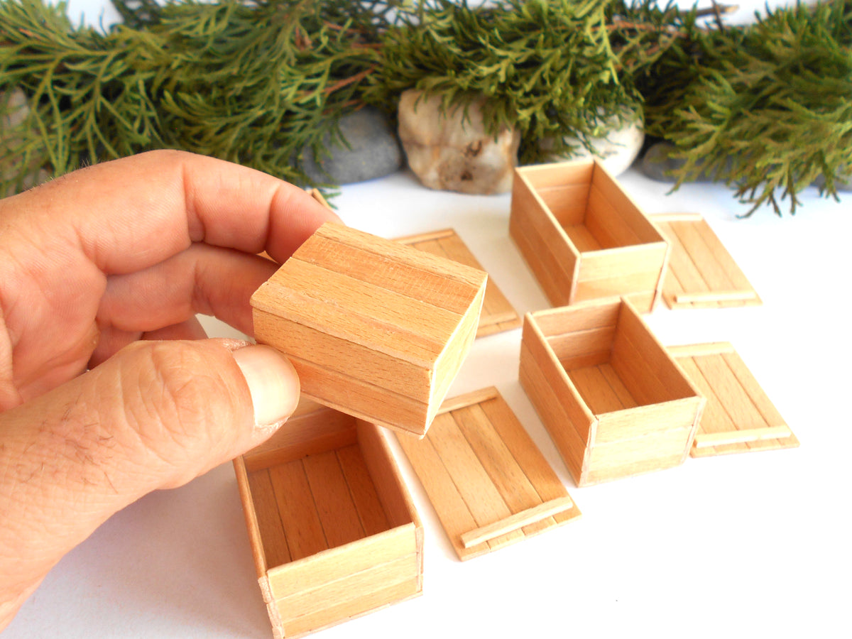 A hand holding a miniature wooden crate box, with several other small wooden storage boxes and lids arranged on a white surface in the background, decorated with green foliage.