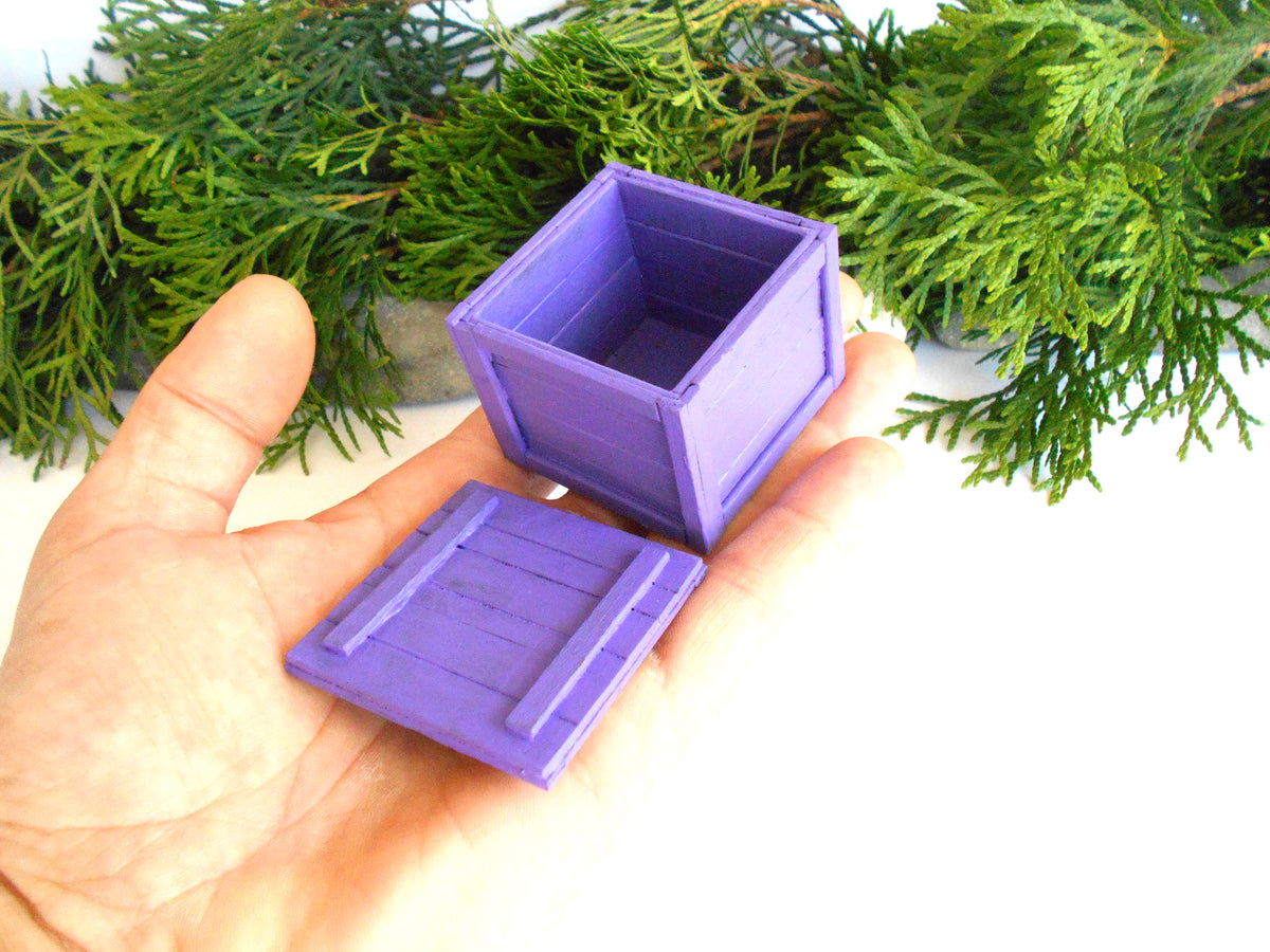A miniature purple plastic box and lid, shown held in a person's hand with green foliage in the background.