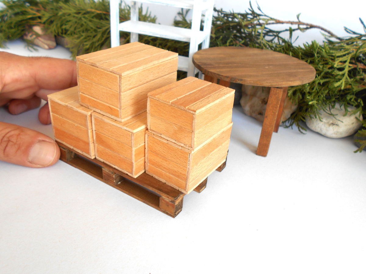 A miniature wooden box stack on a small wooden pallet, held by a human hand to show scale, with a miniature round table and greenery in the background.