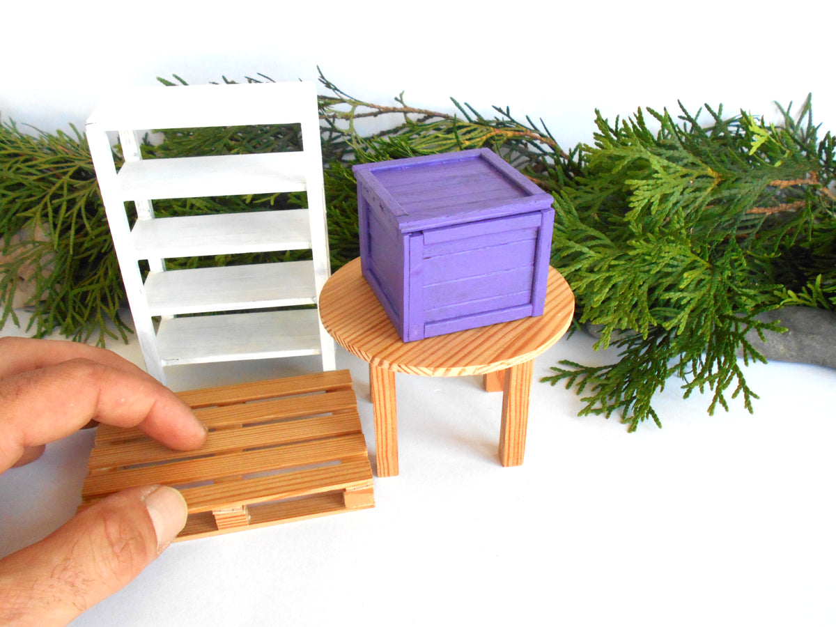 A miniature dollhouse furniture set featuring a purple wooden crate, a small round wooden stool, a white shelving unit, and a wooden pallet, with a hand holding the pallet in the foreground.
