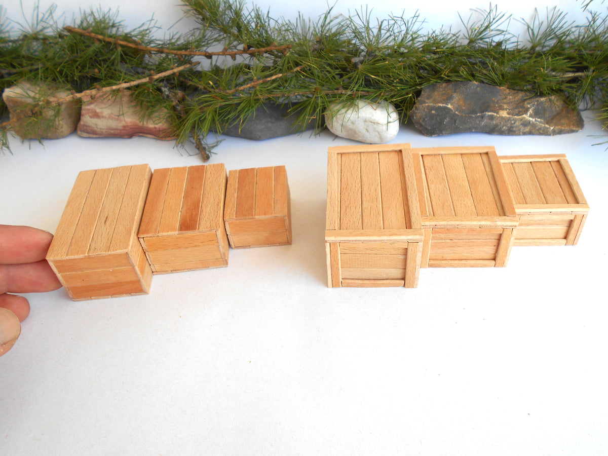 A set of miniature light wood crates, including one being held by a hand to show scale, arranged on a white surface with decorative green pine branches and stones in the background.