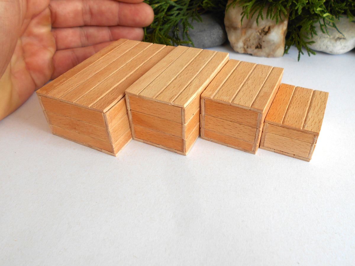 Four miniature wooden boxes or boxes of varying heights arranged in a row on a white surface, with a hand visible on the left for scale and small green plants in the background.