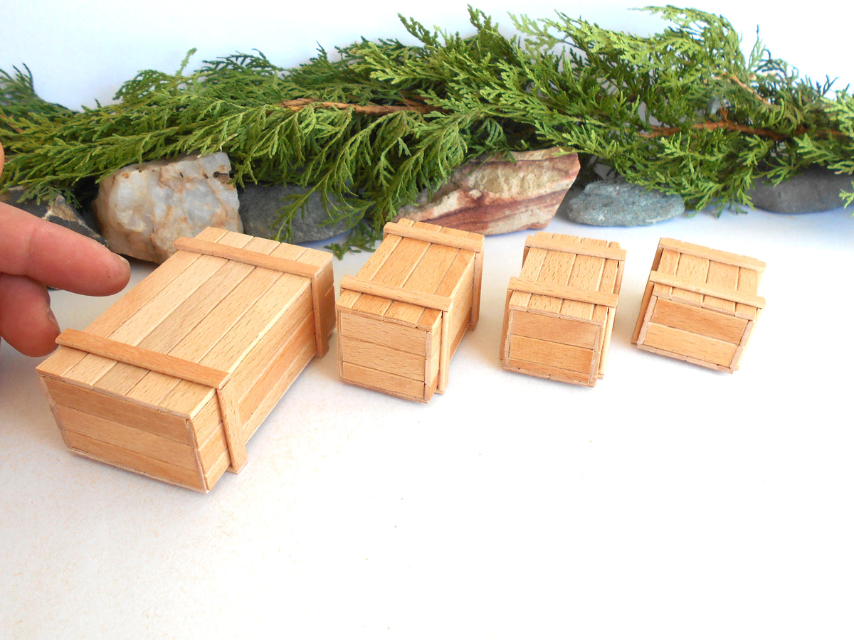 Four miniature light wood crates of different sizes arranged on a white surface, with a hand touching the largest crate and green foliage in the background.