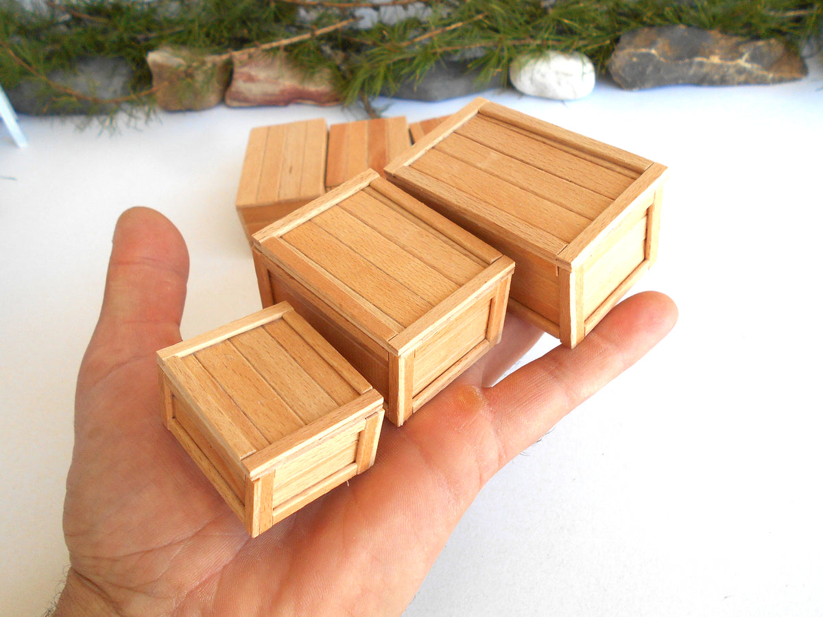 Four miniature wooden crates in different sizes held in a person's hand to show scale, with a white surface and some greenery in the background.