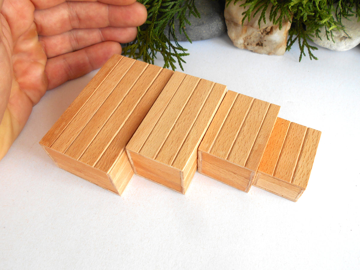 Four small rectangular wooden boxes with grooved sides, arranged in descending size order on a white surface. A hand is visible on the left for scale, with green foliage and rocks in the blurred background.