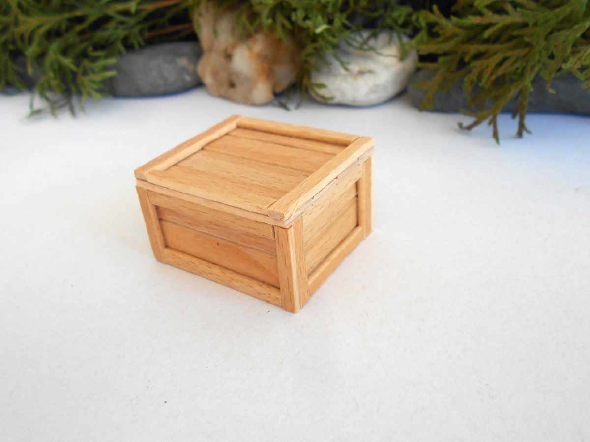 A small, square wooden box with a slatted lid, made from light natural wood, set against a white surface with blurred green foliage in the background.