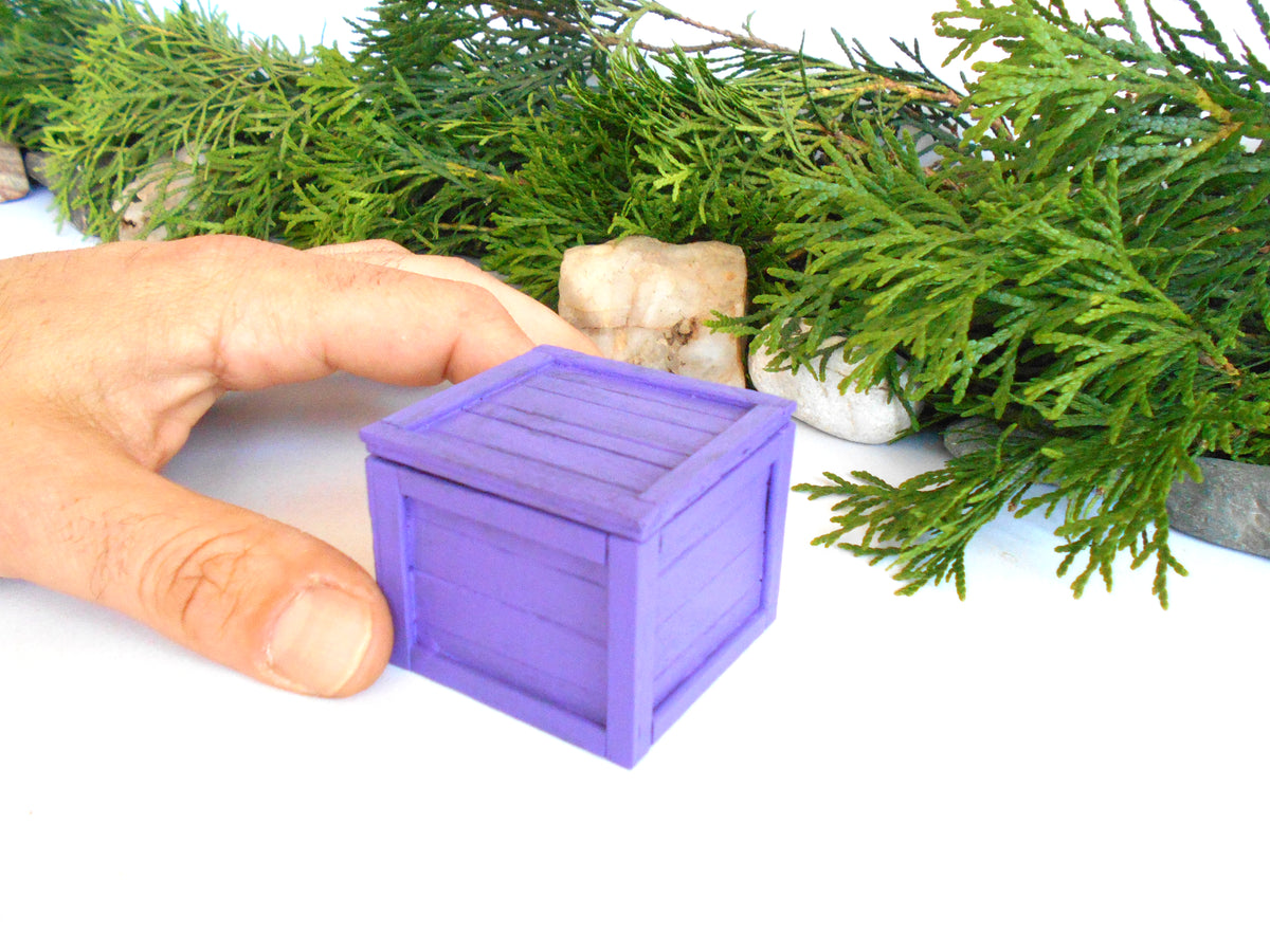 A small purple miniature storage box with a slatted lid, held by a person's hand to show scale, with green pine branches in the background.