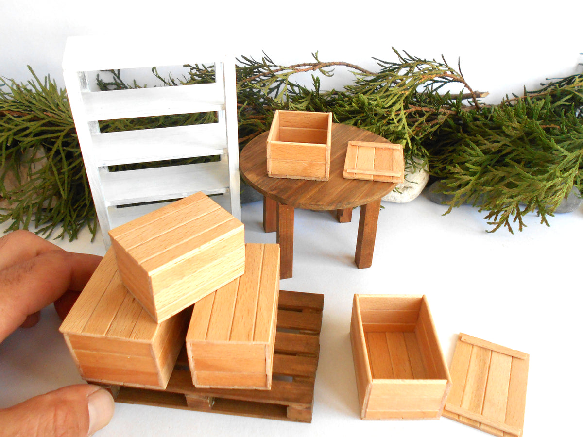 A miniature wooden crate set for dollhouse or model building, featuring various sizes of light brown wooden crates, a small round wooden table, a white slatted shelf, and a miniature wooden pallet. A hand is visible on the left holding one of the crates, set against a white background decorated with green foliage.