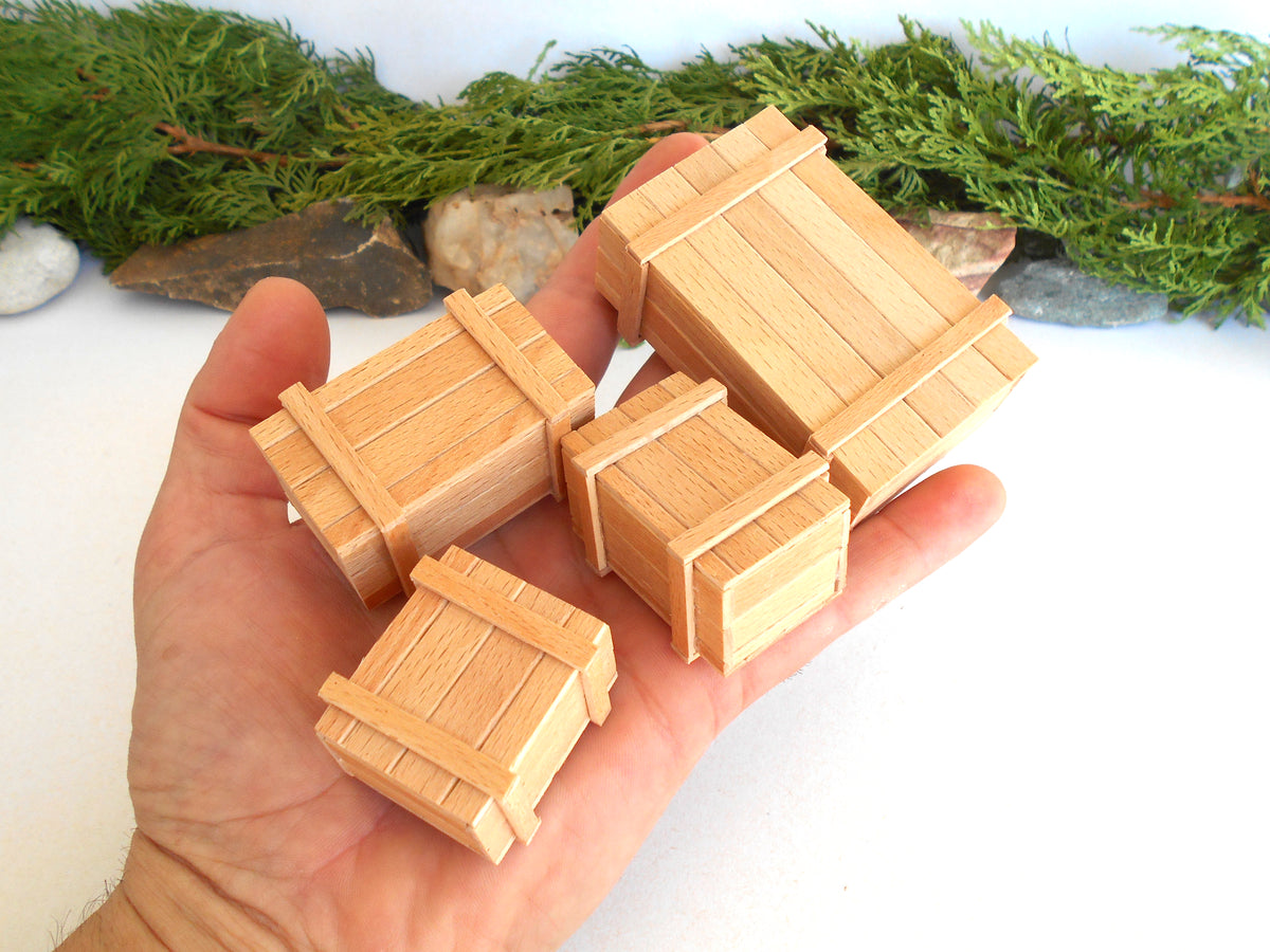 Four miniature wooden crates in different sizes held in a person's hand, with a blurred background of green foliage and rocks.