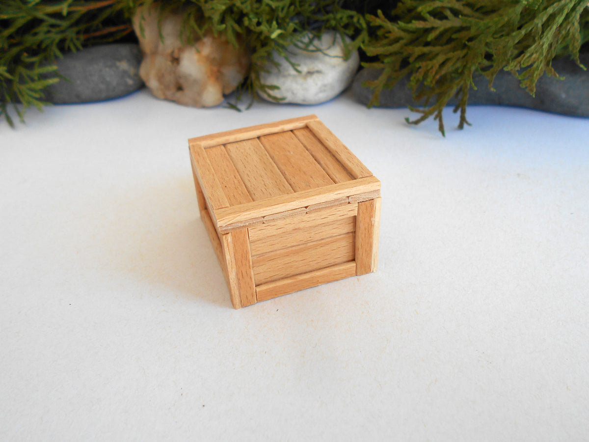 A miniature light wood crate box with a slatted lid, photographed on a white surface with blurred green foliage and stones in the background.