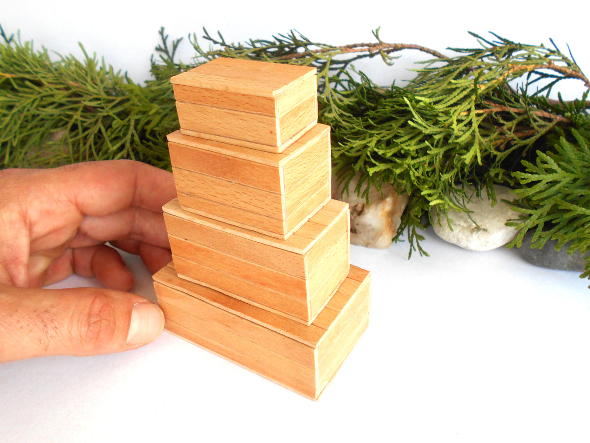 A hand holding a stack of four small, handcrafted wooden boxes with a natural light oak finish, set against a white background with green foliage and small stones.
