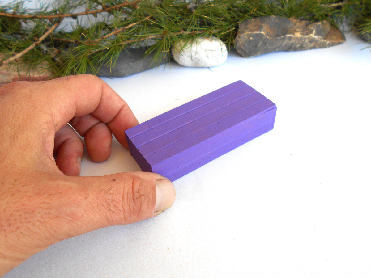 A hand holding a small rectangular purple block with grooved sides, photographed against a white surface with pine branches and stones in the background.