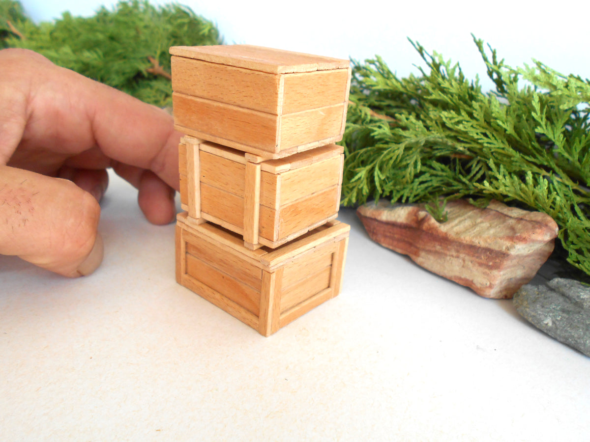 Three miniature light brown wooden boxes stacked vertically, held by a person's hand on a white surface with green foliage and a rock in the background.