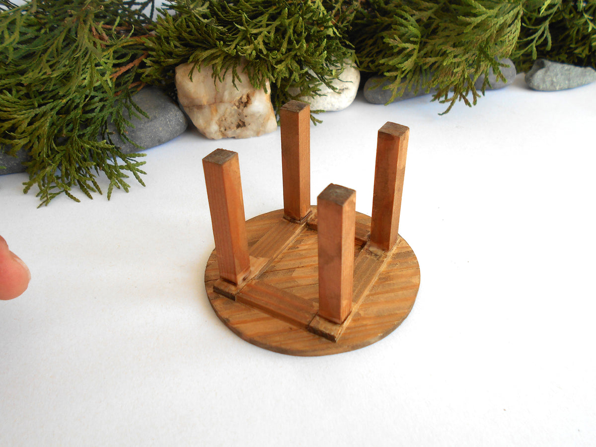 A miniature wooden stool or stand with a round base and four upright legs, photographed on a white surface with decorative greenery and stones in the background.