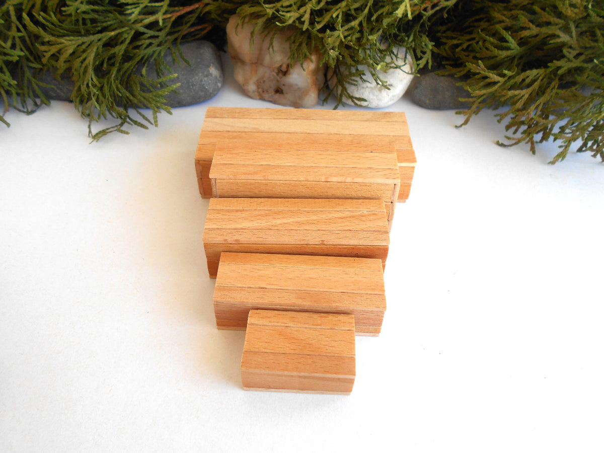 Five natural wood miniature boxes of varying rectangular sizes stacked in a staggered arrangement on a white surface, with green foliage and small stones in the background.
