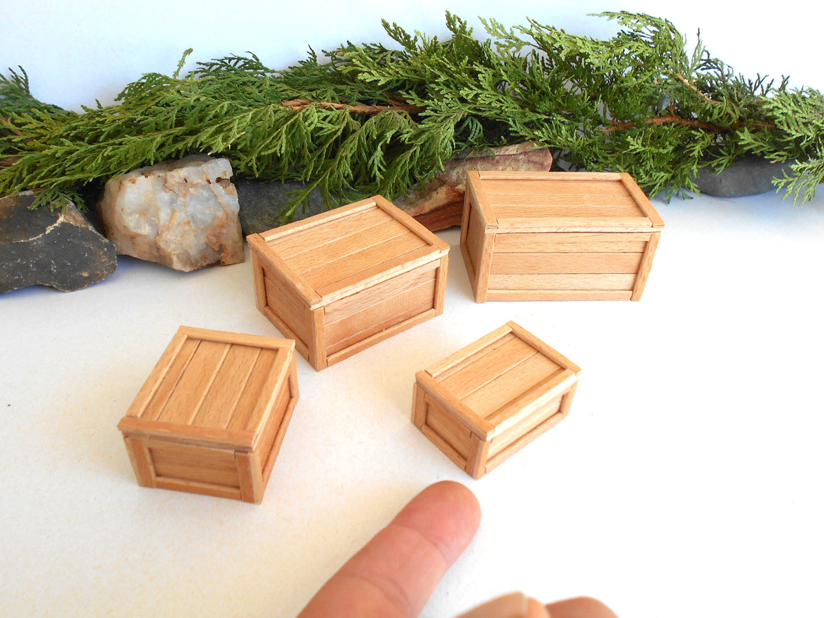 Five miniature light wood crates of different sizes arranged on a white surface with green foliage and rocks in the background. A person's finger is visible at the bottom of the frame for scale.